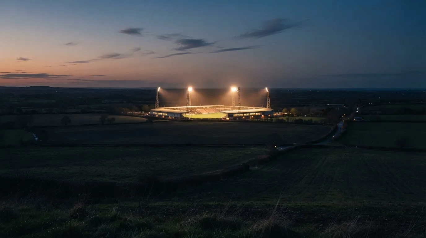 Stadion rozświetlony reflektorami widziany z daleka znad ciemnych pól w pogodny wieczór.