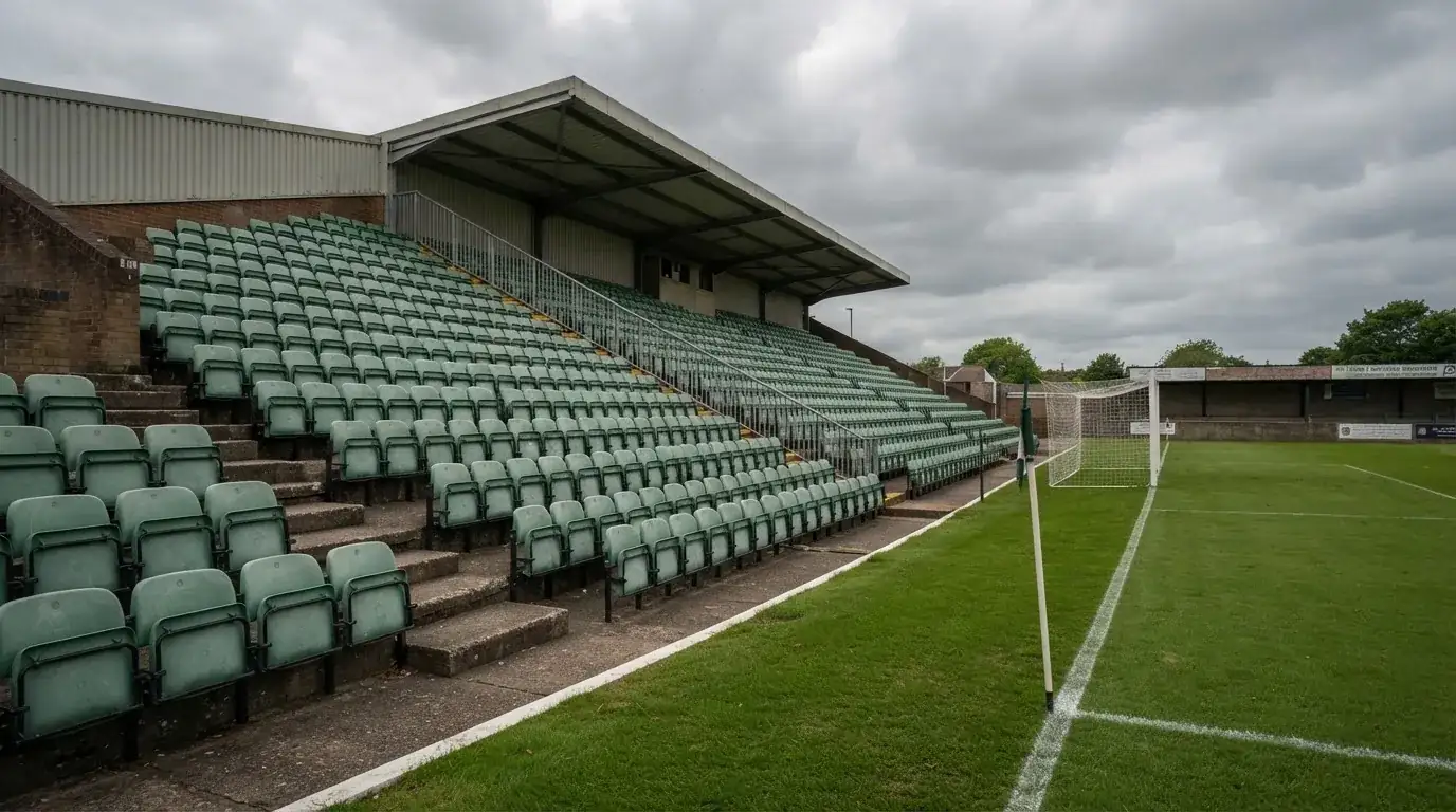 Rzędy pustych plastikowych krzesełek małego stadionu widziane z poziomu murawy pod pochmurnym niebem.