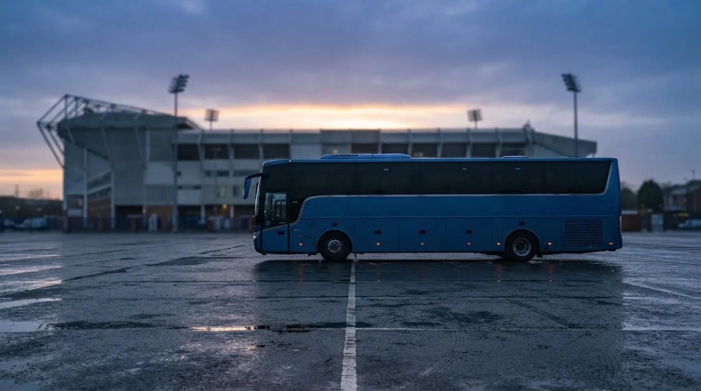 Autokar stoi na parkingu przed stadionem o świcie, rozmyte kontury trybun w tle, pusty plac i mokry asfalt.